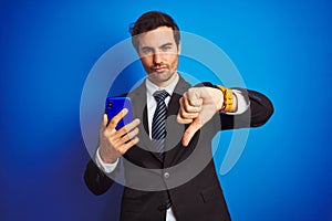 Young handsome businessman using smartphone standing over isolated blue background with angry face, negative sign showing dislike