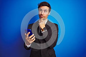 Young handsome businessman using smartphone standing over isolated blue background serious face thinking about question, very