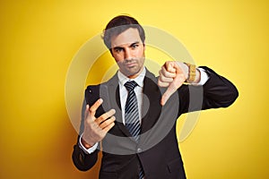 Young handsome businessman using smartphone standing over isolated yellow background with angry face, negative sign showing