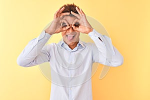 Young handsome businessman wearing elegant shirt over isolated yellow background doing ok gesture like binoculars sticking tongue