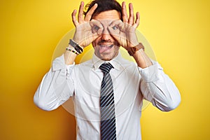 Young handsome businessman wearing elegant shirt and tie over isolated yellow background doing ok gesture like binoculars sticking