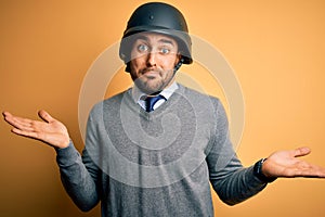 Young handsome businessman wearing military helmet over isolated yellow background clueless and confused expression with arms and