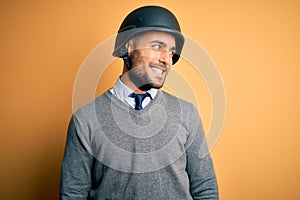 Young handsome businessman wearing military helmet over isolated yellow background looking away to side with smile on face,