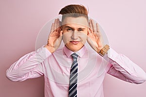 Young handsome businessman wearing shirt and tie standing over isolated pink background Trying to hear both hands on ear gesture,