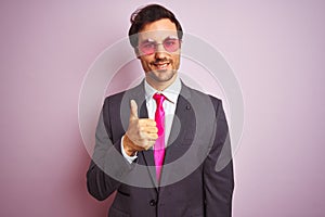 Young handsome businessman wearing suit and sunglasses over isolated pink background doing happy thumbs up gesture with hand