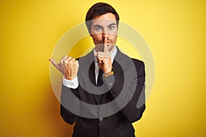 Young handsome businessman wearing suit and tie standing over isolated yellow background asking to be quiet with finger on lips