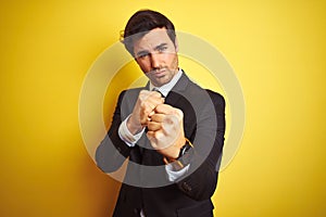 Young handsome businessman wearing suit and tie standing over isolated yellow background Ready to fight with fist defense gesture,