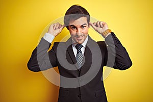 Young handsome businessman wearing suit and tie standing over isolated yellow background Smiling pulling ears with fingers, funny