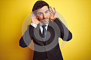 Young handsome businessman wearing suit and tie standing over isolated yellow background Trying to hear both hands on ear gesture,