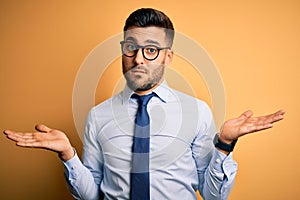Young handsome businessman wearing tie and glasses standing over yellow background clueless and confused with open arms, no idea