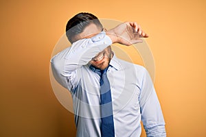 Young handsome businessman wearing tie and glasses standing over yellow background covering eyes with arm smiling cheerful and