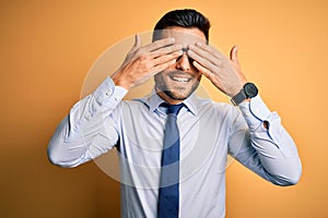 Young handsome businessman wearing tie and glasses standing over yellow background covering eyes with hands smiling cheerful and