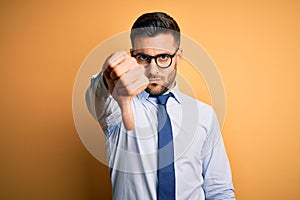 Young handsome businessman wearing tie and glasses standing over yellow background looking unhappy and angry showing rejection and