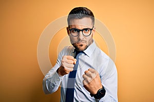 Young handsome businessman wearing tie and glasses standing over yellow background Ready to fight with fist defense gesture, angry