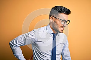 Young handsome businessman wearing tie and glasses standing over yellow background Suffering of backache, touching back with hand,