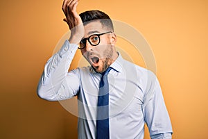 Young handsome businessman wearing tie and glasses standing over yellow background surprised with hand on head for mistake,