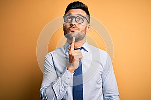 Young handsome businessman wearing tie and glasses standing over yellow background Thinking concentrated about doubt with finger