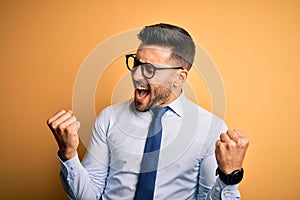 Young handsome businessman wearing tie and glasses standing over yellow background very happy and excited doing winner gesture