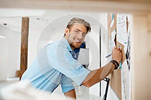 Young handsome businessman writing on the paper pinned to corkboard, looking in camera smiling. Office background.