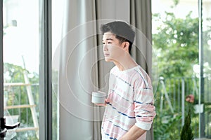 Young handsome carefree man near modern full length window enjoying a cup of coffee while looking outside