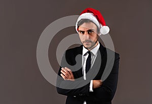 Young handsome caucasian guy in business suit and Santa hats stands on gray background in studio with serious face folded arms on