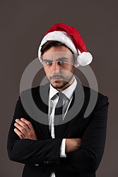 Young handsome caucasian guy in business suit and Santa hats stands on gray background in studio with serious face folded arms on