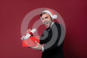 Young handsome caucasian guy in business suit and Santa hats stands on red background in studio and teeth smilie Holding red gift