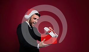 Young handsome caucasian guy in business suit and Santa hats stands on red background in studio and teeth smilie Holding red gift