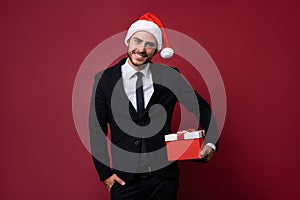 Young handsome caucasian guy in business suit and Santa hats stands on red background in studio and teeth smilie Holding red gift