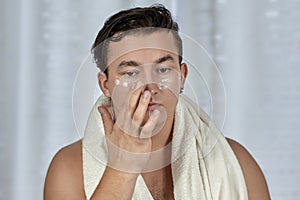 Young handsome caucasian man applying cream under the eyes, towel on shoulders. Caring face, metrosexual daily routine in the bath photo