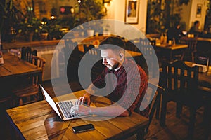 A young handsome Caucasian man with beard and toothy smile in a red checkered shirt is working behind a gray laptop sitting at a w