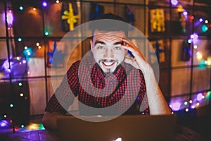 A young handsome Caucasian man with beard and toothy smile in a red checkered shirt is working behind a gray laptop sitting at a w
