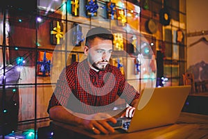 A young handsome Caucasian man with beard and toothy smile in a red checkered shirt is working behind a gray laptop