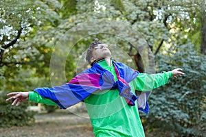 Young handsome caucasian man with bright freaky sportswear 70s style stands at a morning park. Stick in the mouth, golden glasses,