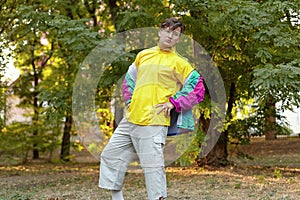 Young handsome caucasian man with bright freaky sportswear 70s style stands at a morning park. Stick in the mouth, golden glasses,