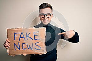 Young handsome caucasian man holding protest carboard for desinformation and fake news with surprise face pointing finger to