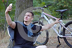 Young handsome caucasian man makes selfie on mobile phone, sitting on the ground close to the big tree at the abandoned park. Bicy