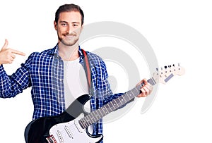 Young handsome caucasian man playing electric guitar pointing finger to one self smiling happy and proud