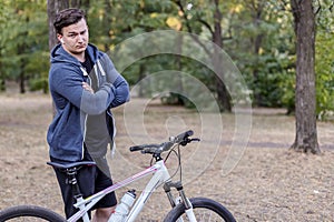Young handsome caucasian man stands hands crossed with bicycle frowning and serious. White wireless earphones, casual sportswear.