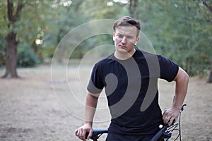Young handsome caucasian man stands with his bicycle at the middle of park or forest. Very serious and thoughful, frowns. White ea