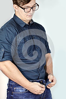 Young handsome caucasian man unzipps blue jeans. Man torso, fingerring, blue shirt, glasses.