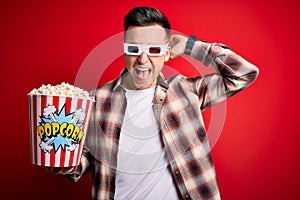 Young handsome caucasian man wearing 3d movie glasses and eating popcorn Crazy and scared with hands on head, afraid and surprised