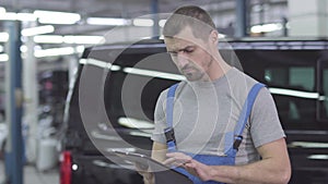 Young handsome Caucasian man in workwear standing with tablet at the background of black car. Serious male auto mechanic