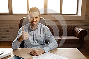 Young handsome cheerful businessman sit on table and pose in his own office. He hold big thumb up and smile. Nice happy