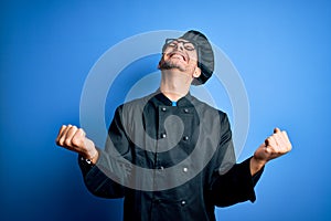 Young handsome chef man wearing cooker uniform and hat over isolated blue background very happy and excited doing winner gesture