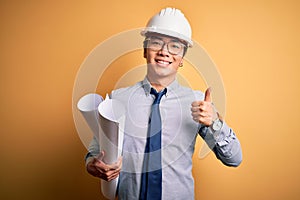 Young handsome chinese architect man wearing safety helmet holding blueprints happy with big smile doing ok sign, thumb up with