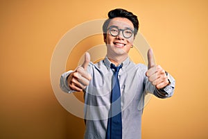 Young handsome chinese businessman wearing glasses and tie over yellow background approving doing positive gesture with hand,