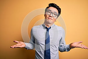 Young handsome chinese businessman wearing glasses and tie over yellow background clueless and confused expression with arms and
