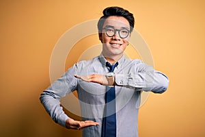 Young handsome chinese businessman wearing glasses and tie over yellow background gesturing with hands showing big and large size