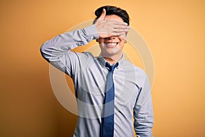 Young handsome chinese businessman wearing glasses and tie over yellow background smiling and laughing with hand on face covering